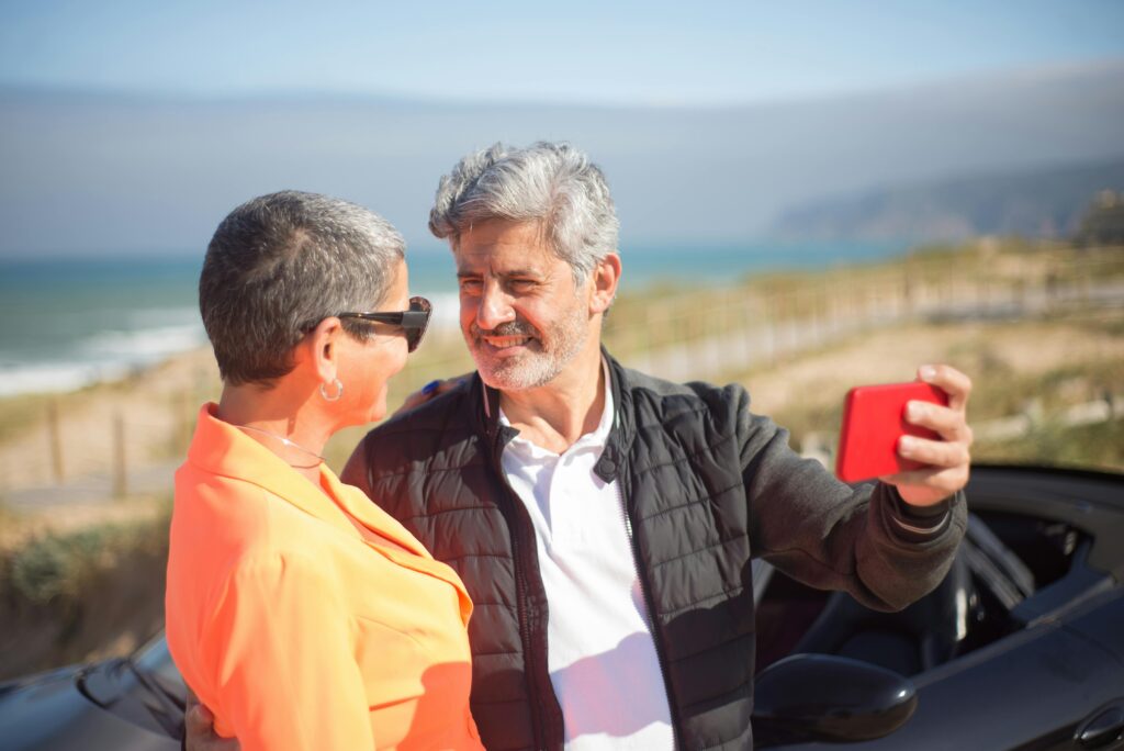 A senior couple enjoying a sunny day at the beach while taking a selfie with a smartphone.
