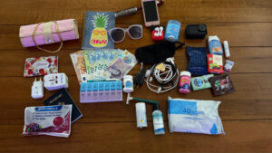 Carry-on travel essentials laid out on a table, including passport, medications, chargers, toiletries, snacks, sunglasses, and travel bag.